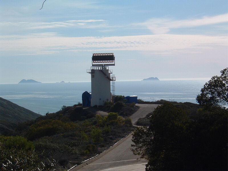 The low dielectric large antenna test fixture is shown installed at its permanent location. It is positioned on top of a white tower with stairs. The tower is located on a hillside overlooking the ocean on a cloudy day.